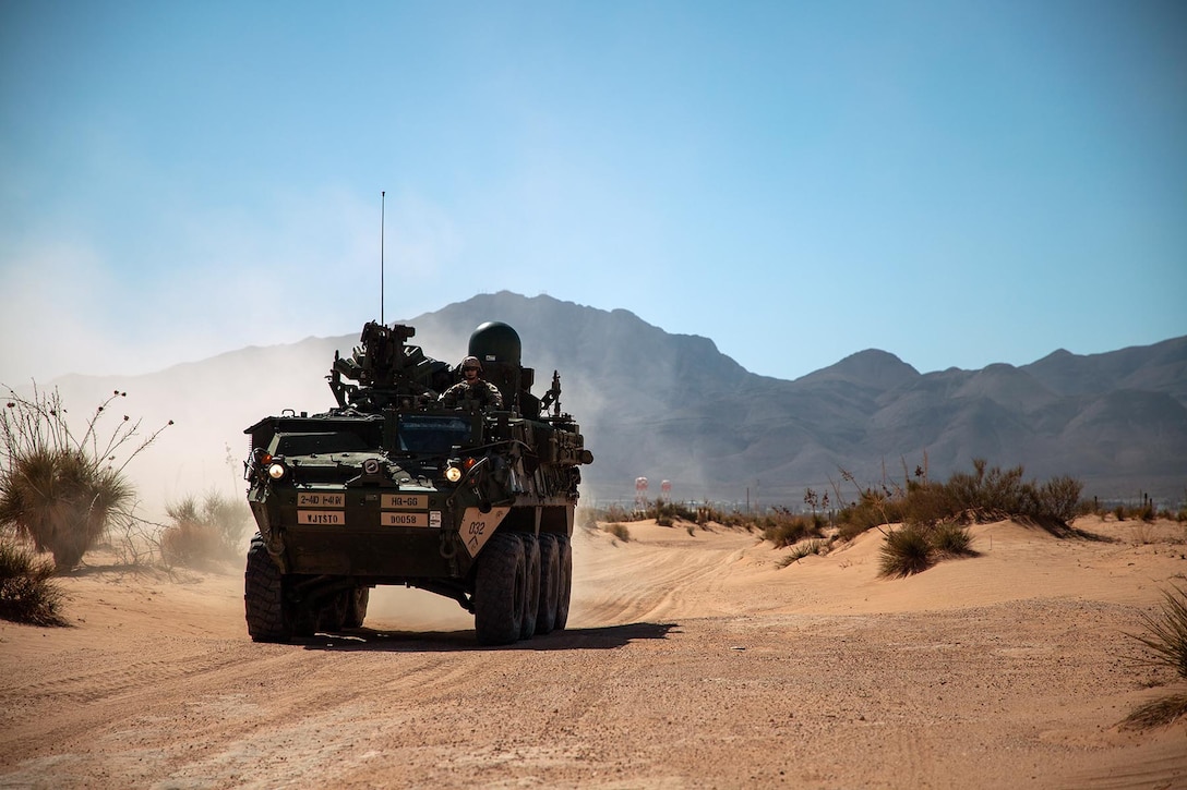 Soldiers ride in an armored vehicle on a dirt road with mountains in the distance under a blue sky.