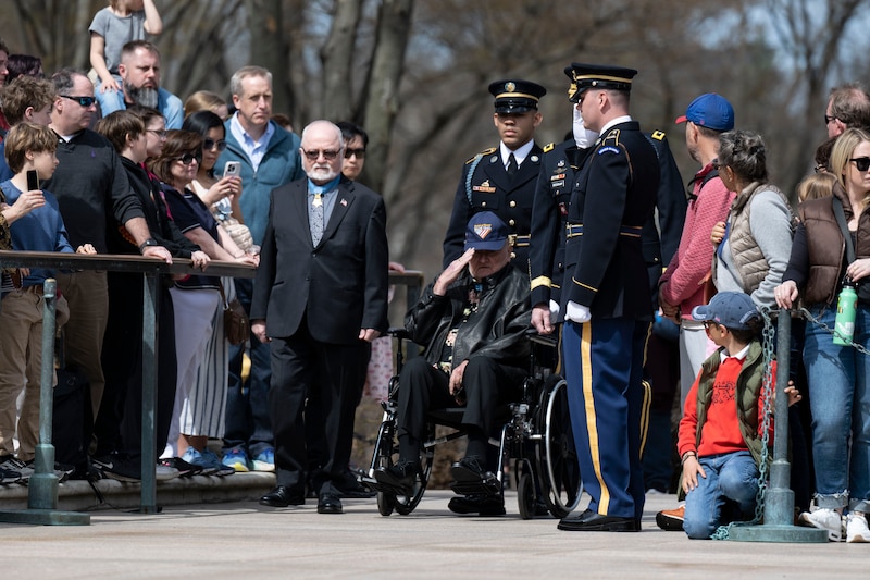 Two veterans are escorted by service members in uniform while crowds to their left and right look on.