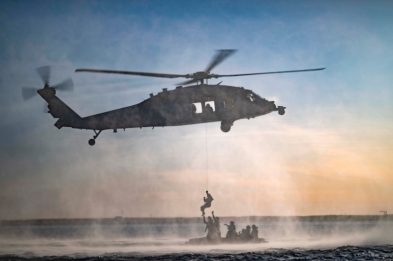 Several sailors in a small craft reach up to a sailor hanging from the rope attached to a helicopter above.