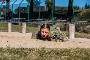 A soldier crawls on sand under barbed wire on a sunny day.