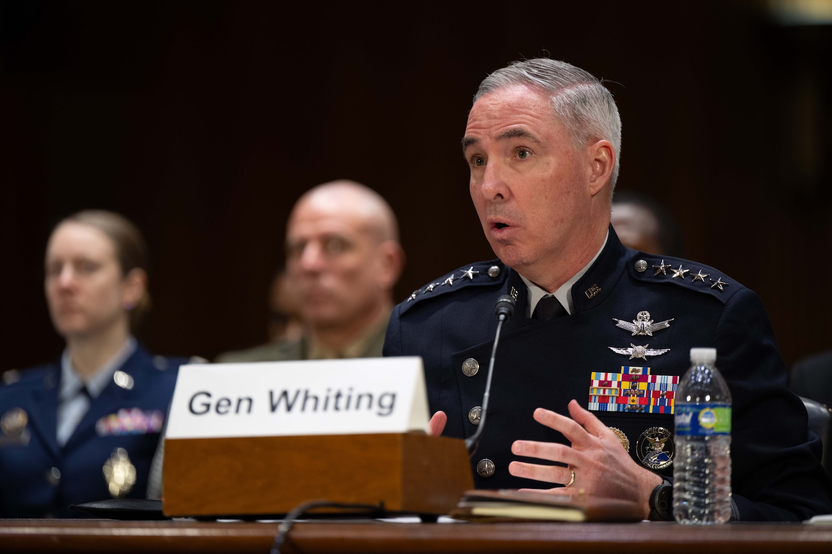 A man in a military suit speaks into a microphone behind a nameplate while seating down.