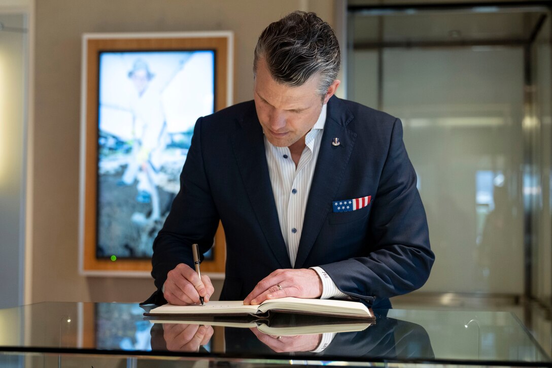 Defense Secretary Pete Hegseth stands and signs a book on a table