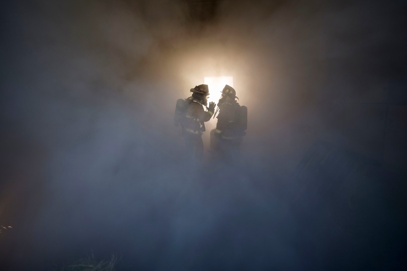 firefighters stand in the midst of a room full of smoke.