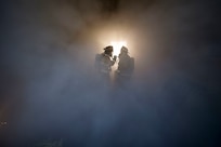 firefighters stand in the midst of a room full of smoke.