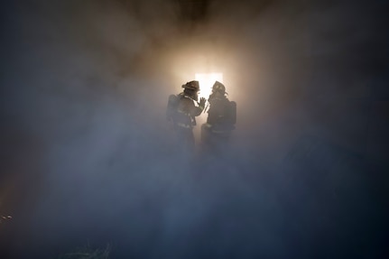 firefighters stand in the midst of a room full of smoke.