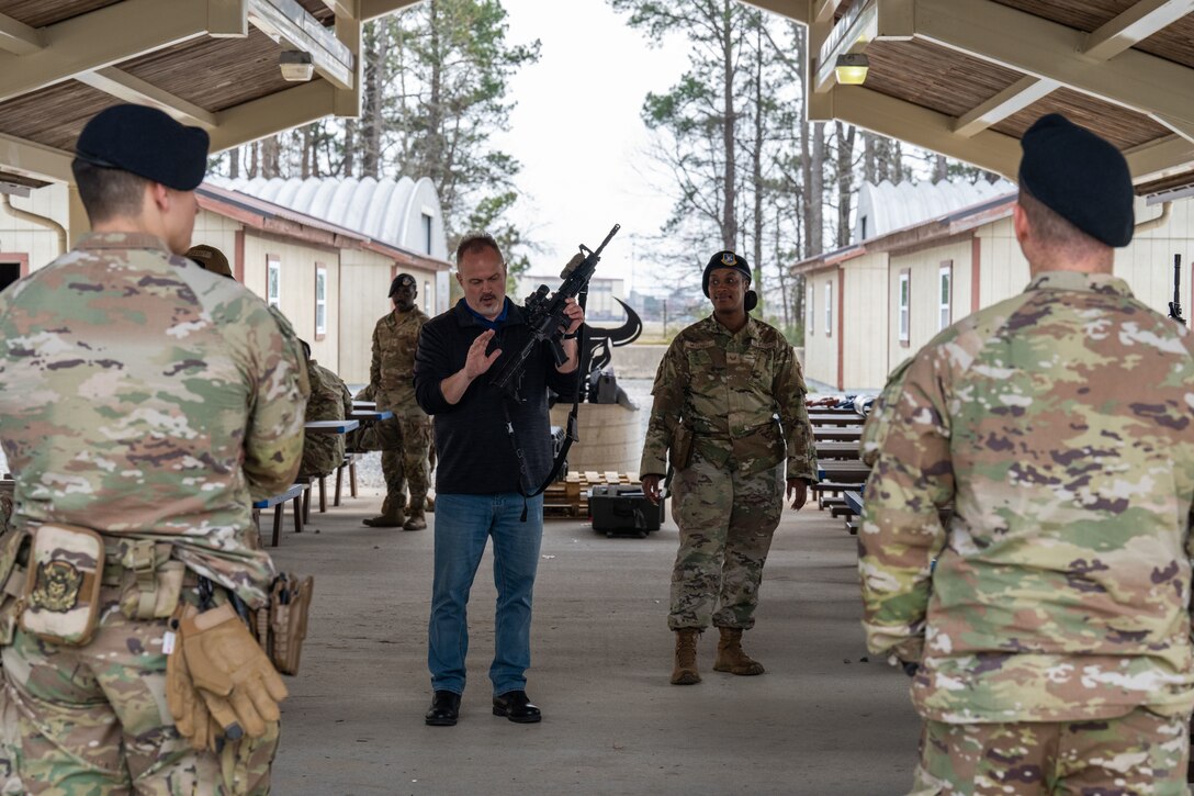 A man holds a rifle pointing out different items while uniformed members watch.
