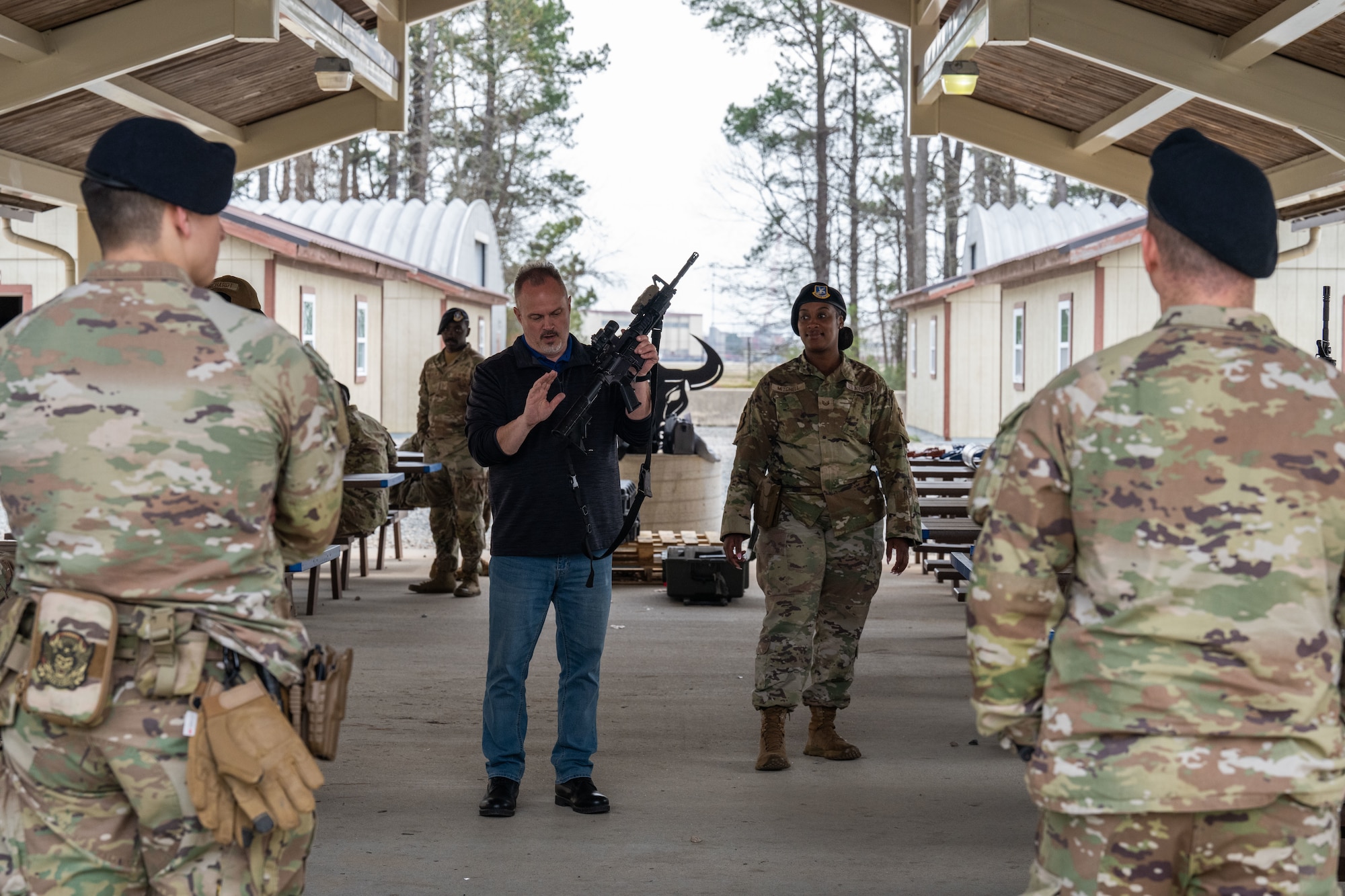 A man holds a rifle pointing out different items while uniformed members watch.