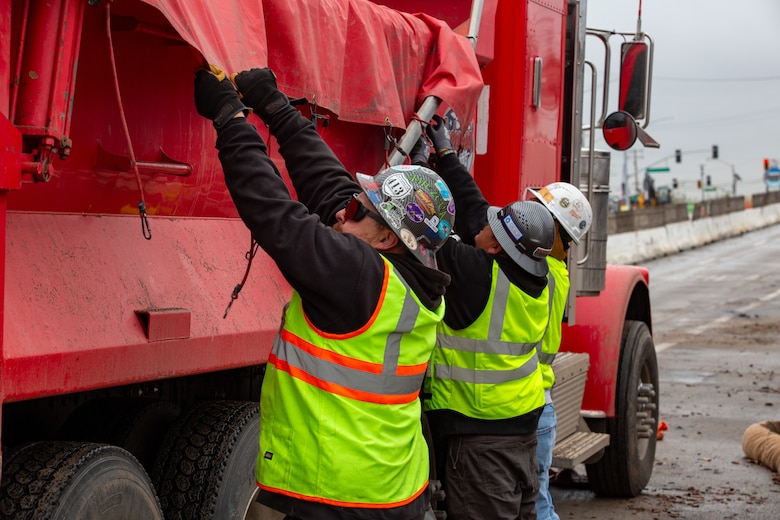 Two men with hard hats and vests put wrapping on debris in a truck