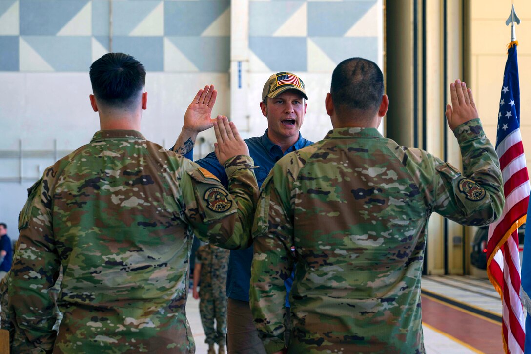 A man wearing civilian clothes and a hat raises his right hand and swears in a pair of soldiers who also raise their right hands.