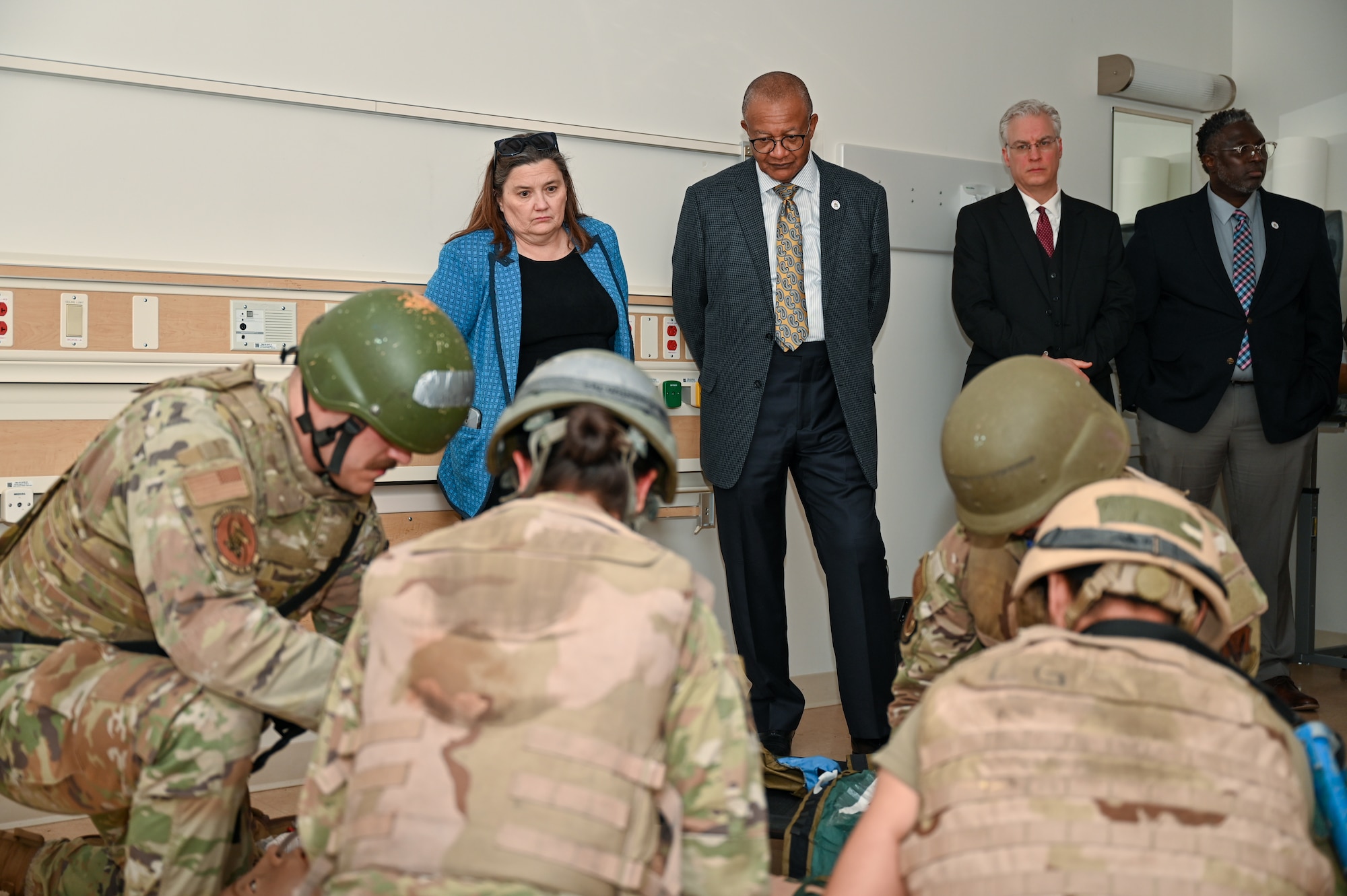 Four military members in the foreground wearing OCPs and helmets while 4 city officials stand behind watching as they work on a fake patient on the ground.