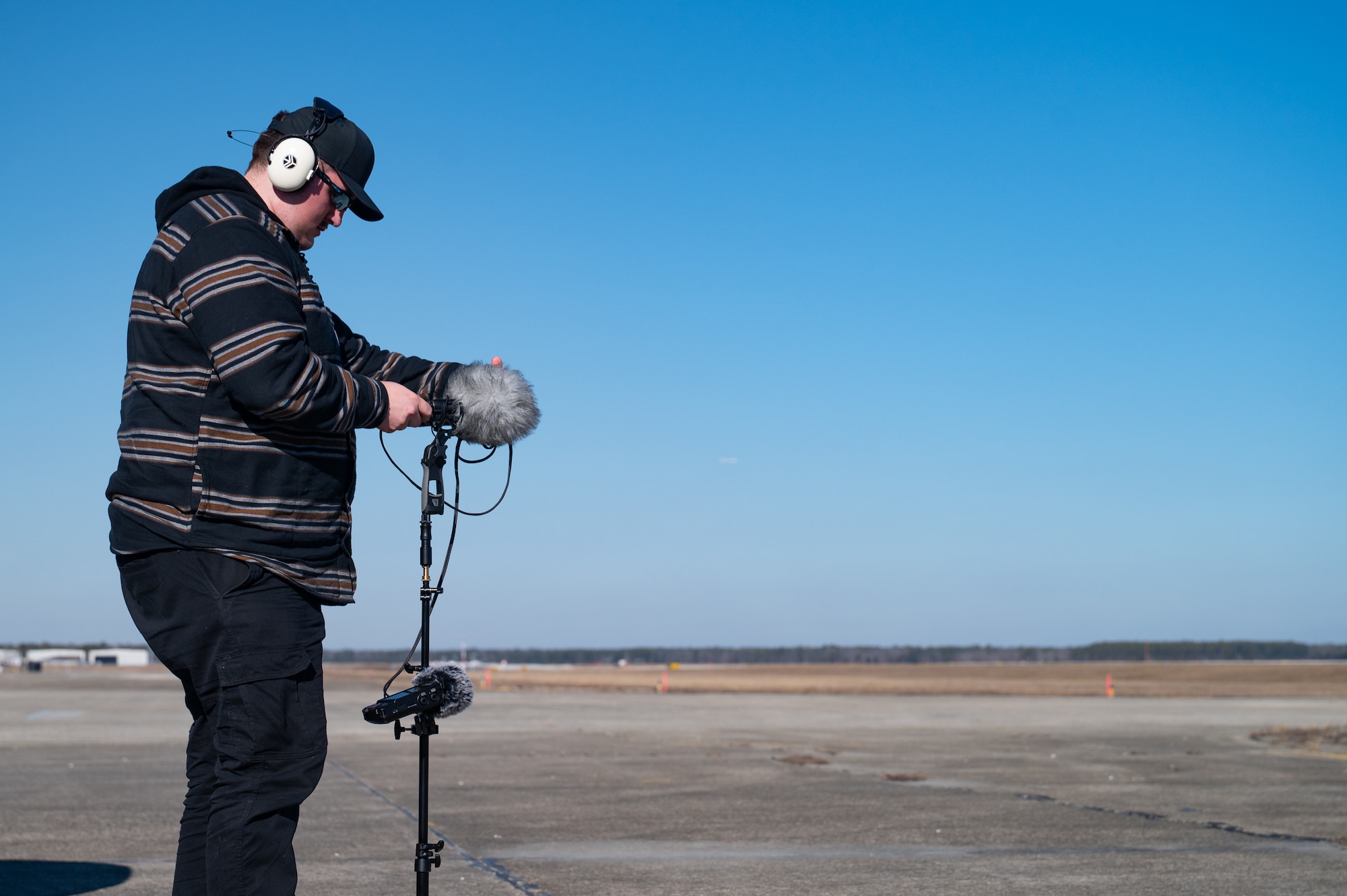 A man stands on a flightline while adjusting audio recording equipment.