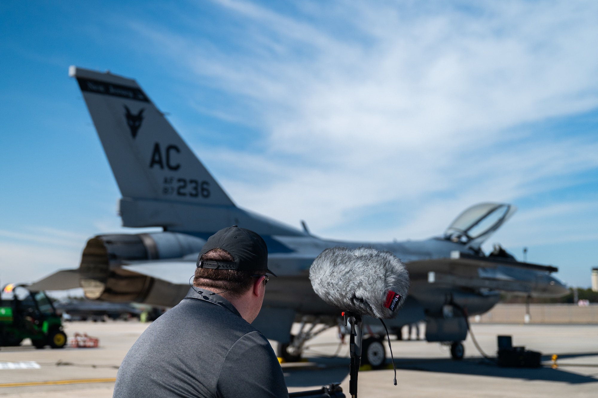 A man and a microphone stand in the foreground of an F-16 fighter jet.