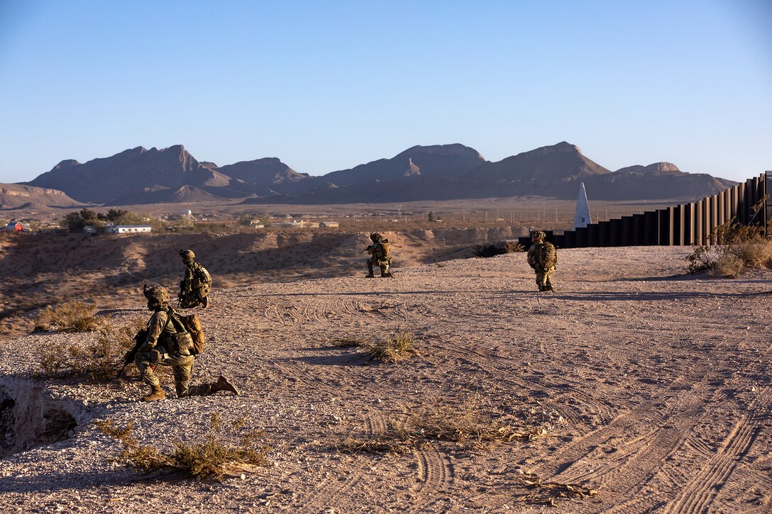 Soldiers in tactical gear kneel in the dirt near a tall wall with mountains in the background.