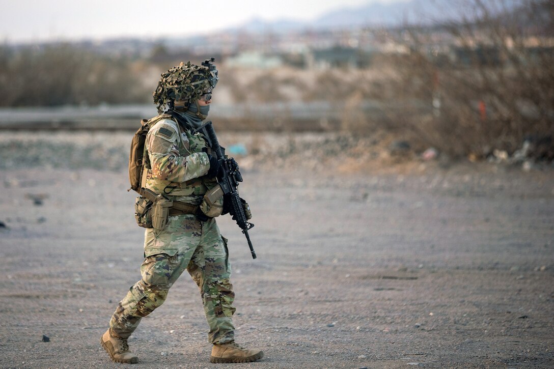 A soldier in tactical gear walk on a dirt road.