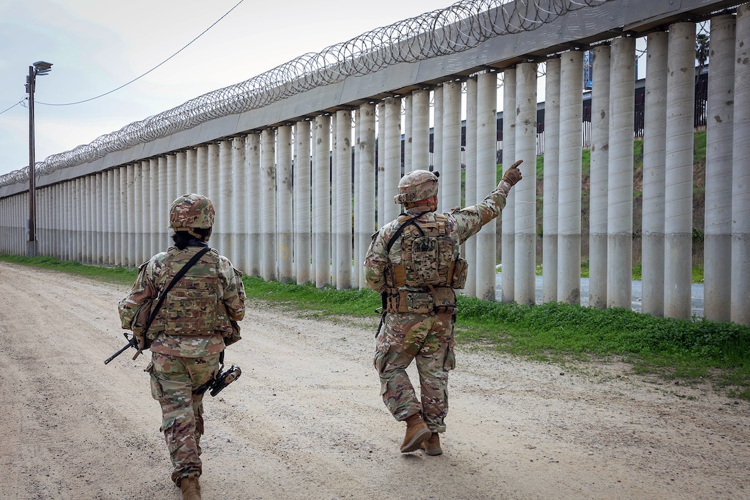 Two soldiers in tactical gear walk near a border wall.
