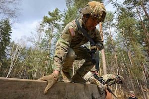 Soldiers climb over a wooden barrier in a forest setting.