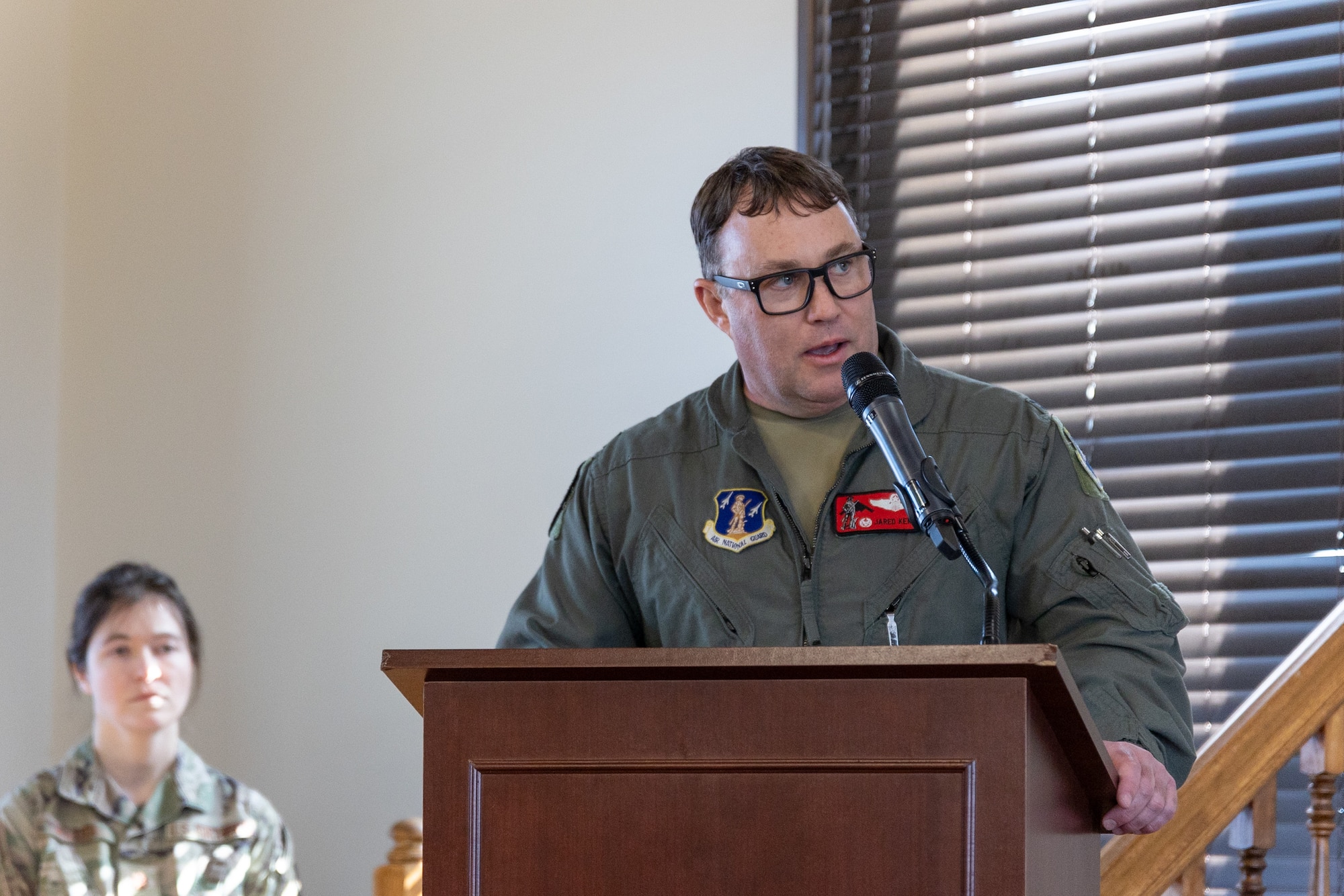 A U.S. Air National Guard Airman speaks from a lectern during an official ceremony. The Airman is wearing a military flight suit.