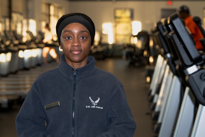 African- American female wearing a blue sweater with the white Air Force logo standing in front of a row of treadmills for a portrait shot.
