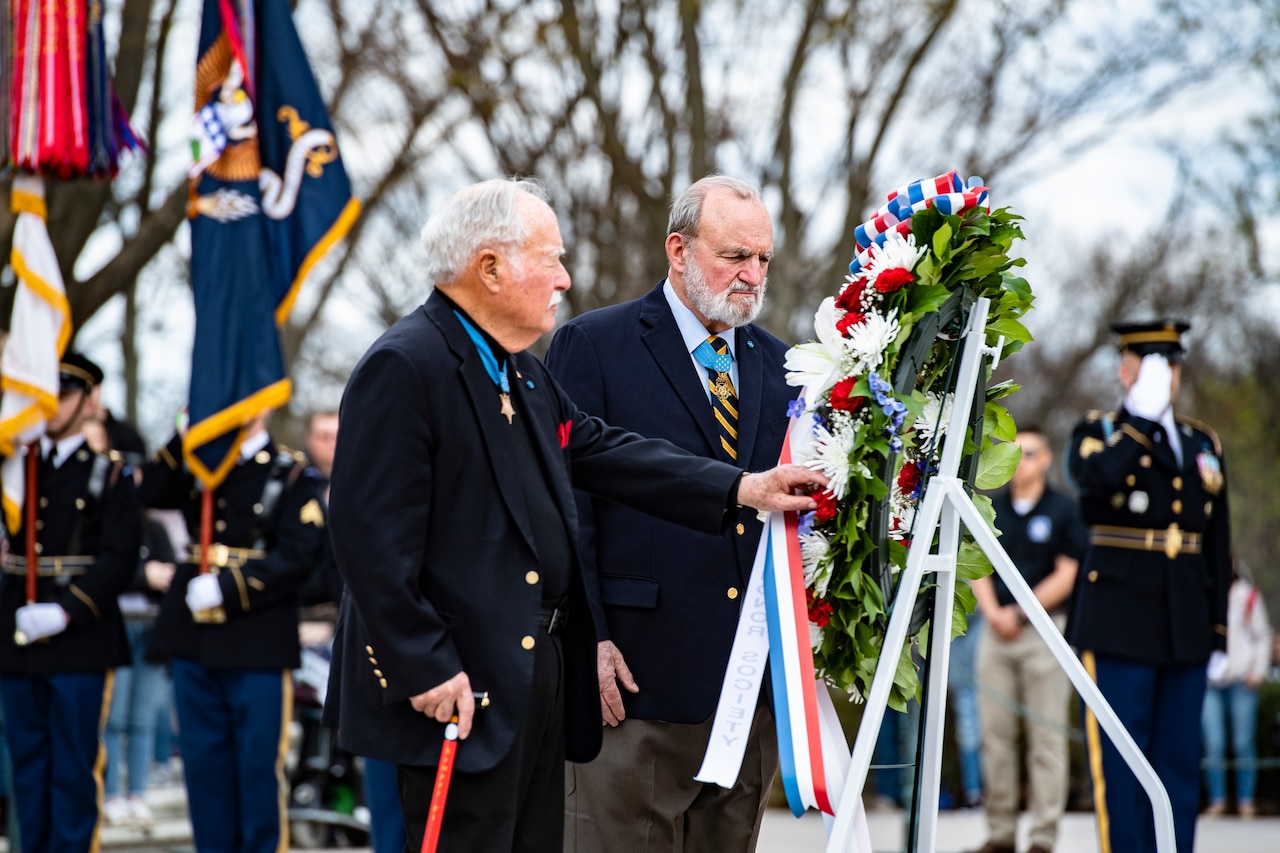 Two men in business attire adjust a wreath on a stand while men stand behind.