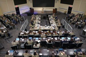 A large group of military members sit in a presentation hall listening to a single speaker.