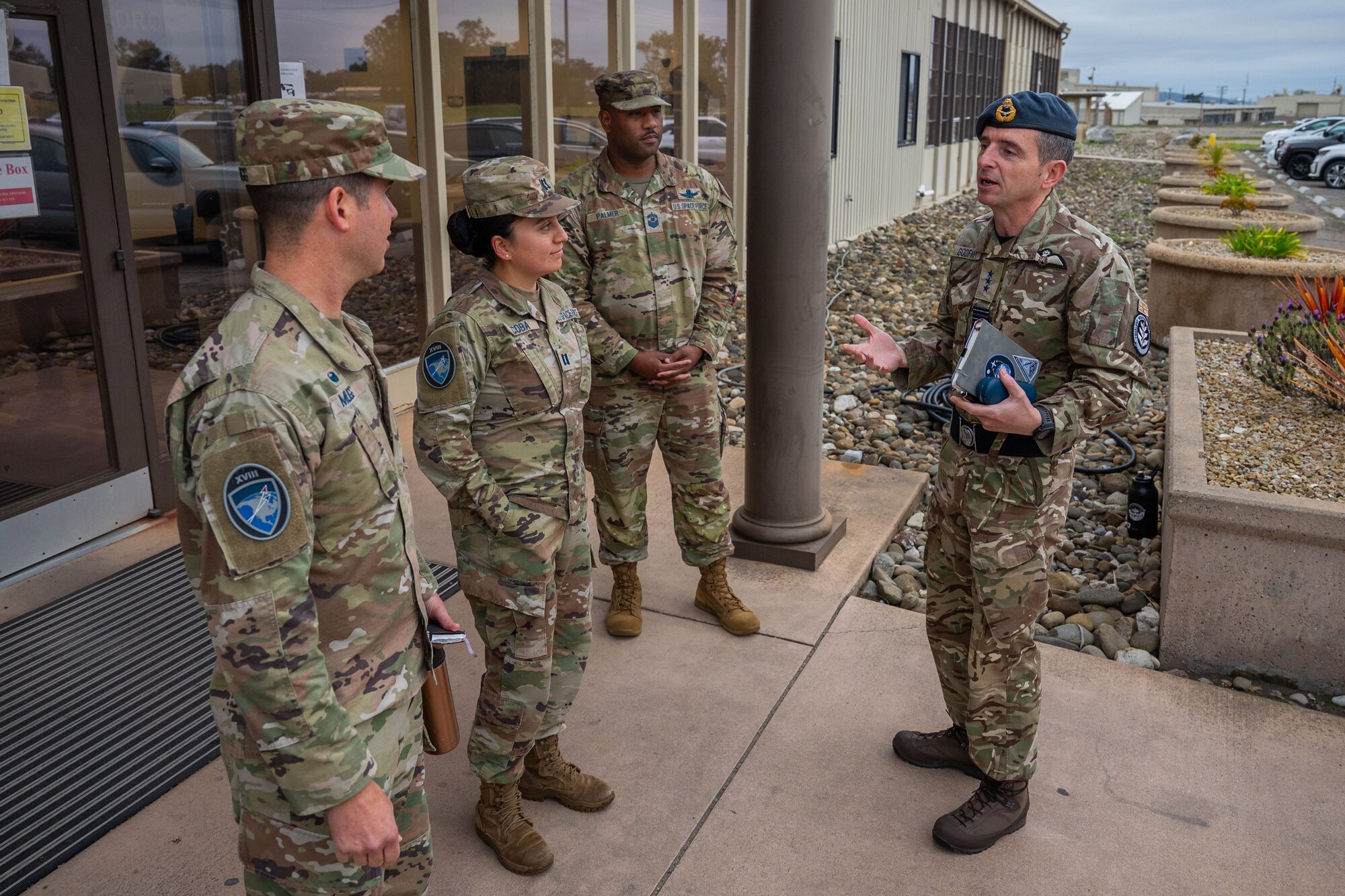 Four military members in uniform discuss topics outside the entrance of a building.