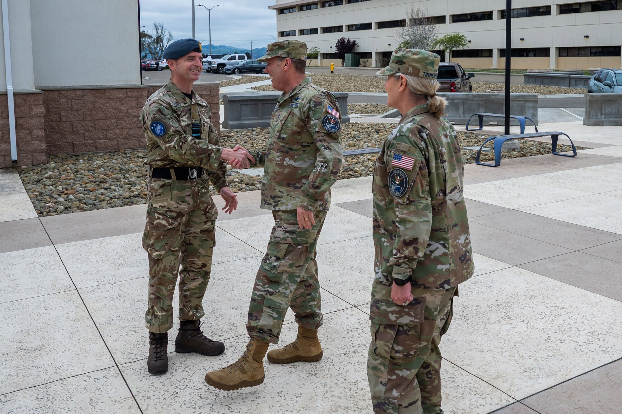 Two military members shake hands in a concrete courtyard outside with another member waiting to be greeted.