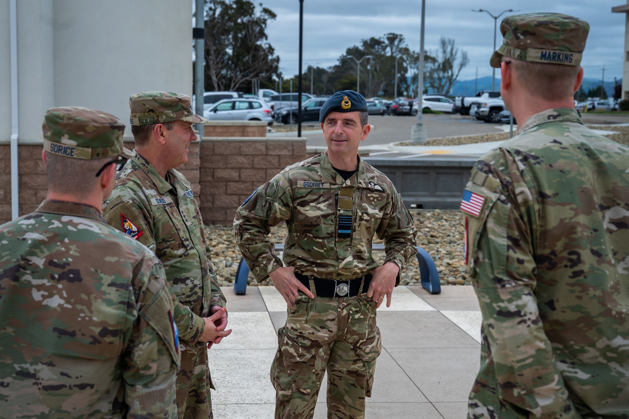 Four military members in uniform stand in a concrete courtyard outside discussing topics with one another.