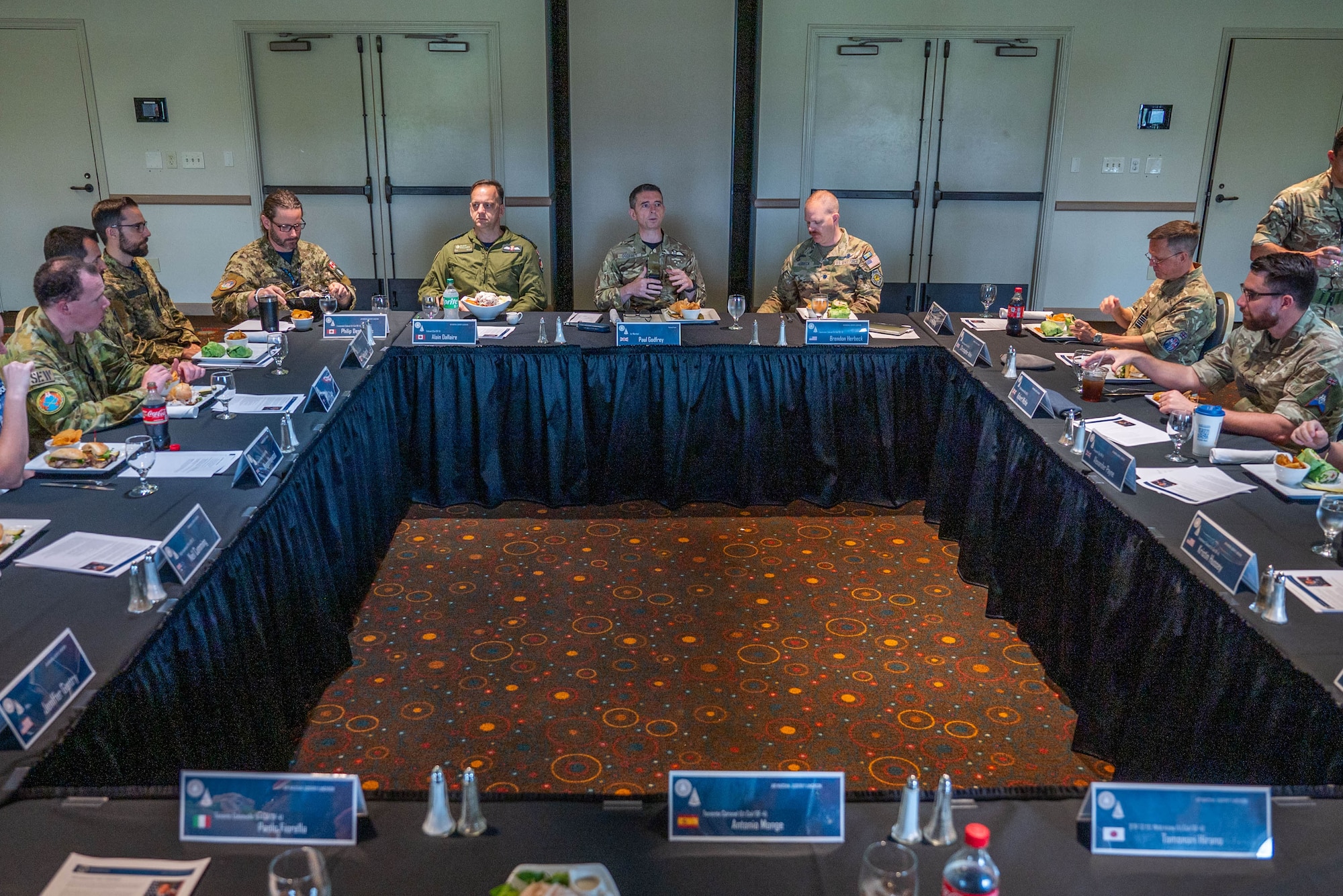 Military members in uniform from multiple nations sit at a square lunch table discussing topics while they eat.