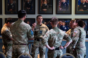 Six military members discuss topics in a circle inside of a room with portraits hanging on the wall.
