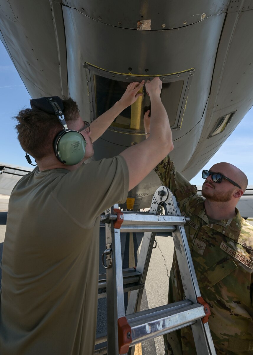 Two men in military uniforms screw a window panel back on a military aircraft.