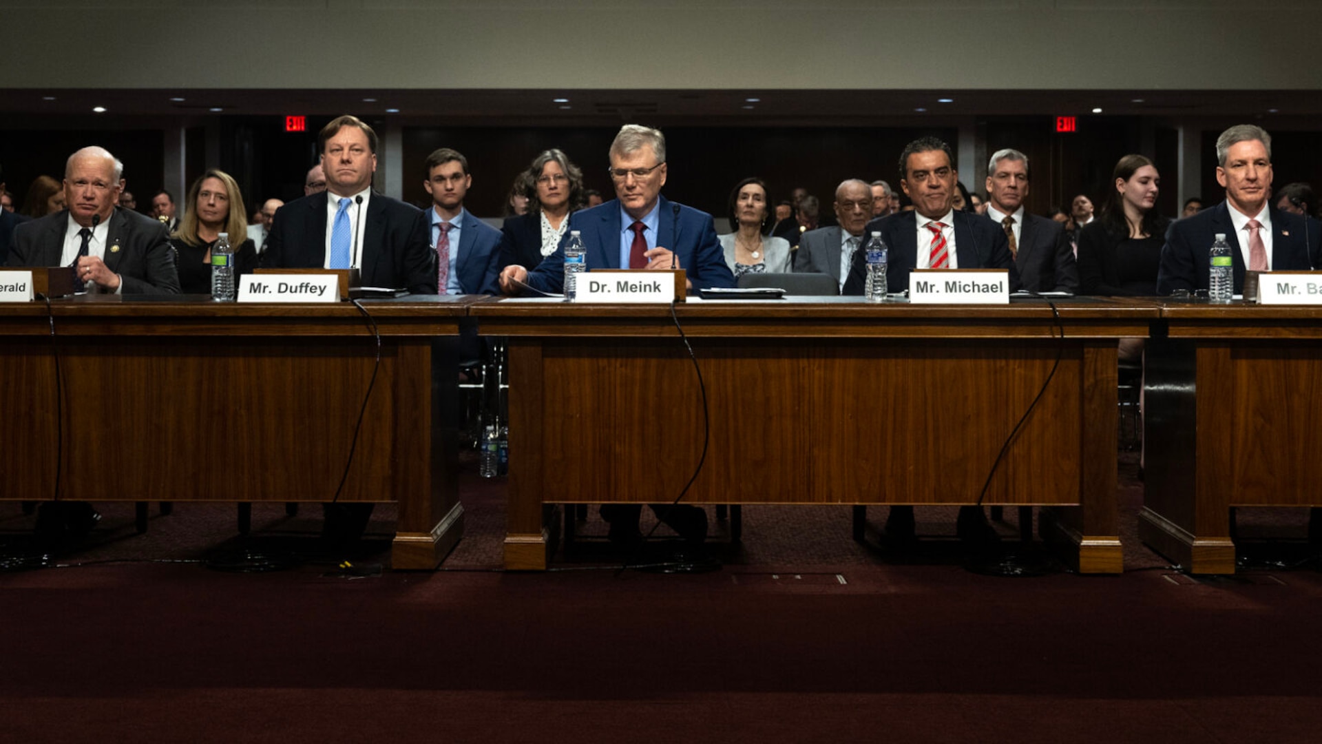 Four men in business attire sit alongside each other at a series of desks.