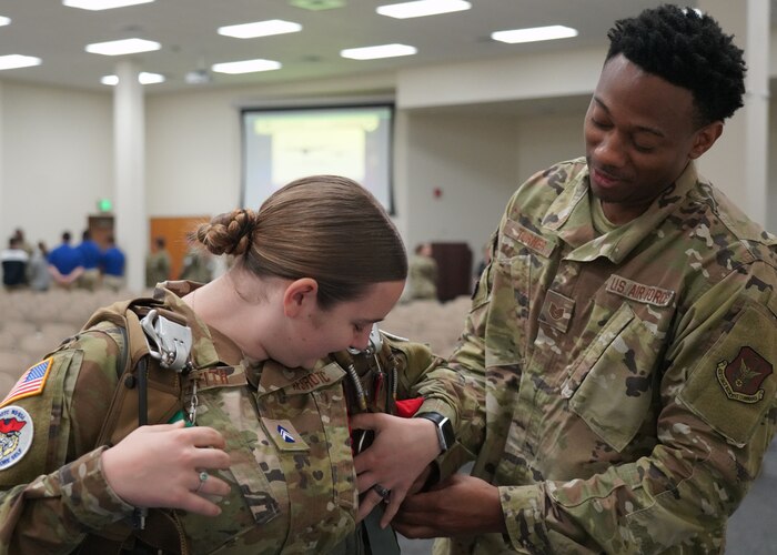 Male Airmen demonstrates to female JROTC cadet how to don a parachute.