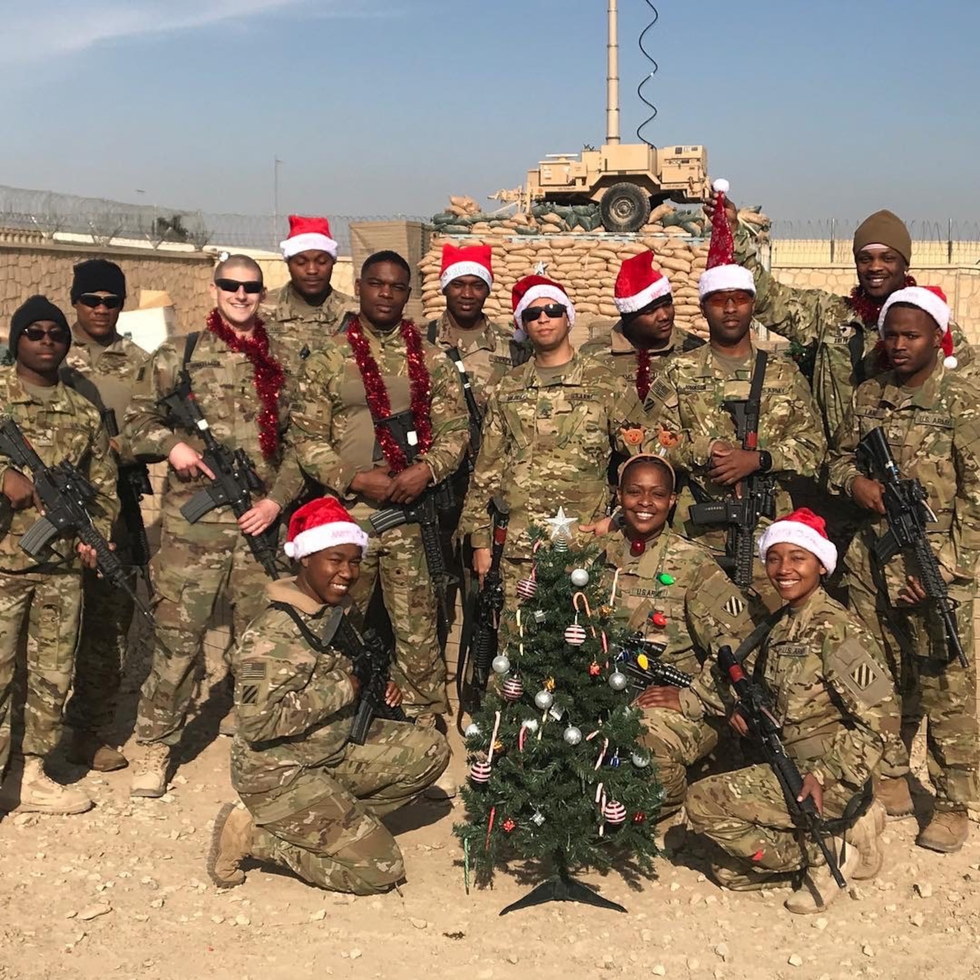 Group of U.S. Army Soldiers in uniform - some with Santa hats on - pose around a small Christmas tree during a deployment.