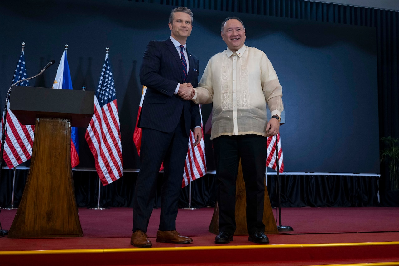 Defense Secretary Pete Hegseth and another official shake hands on a stage with flags behind them.