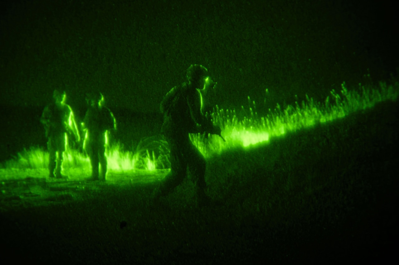 Three soldiers in tactical gear move through a field in the dark illuminated by green light as seen through a night vision lens.