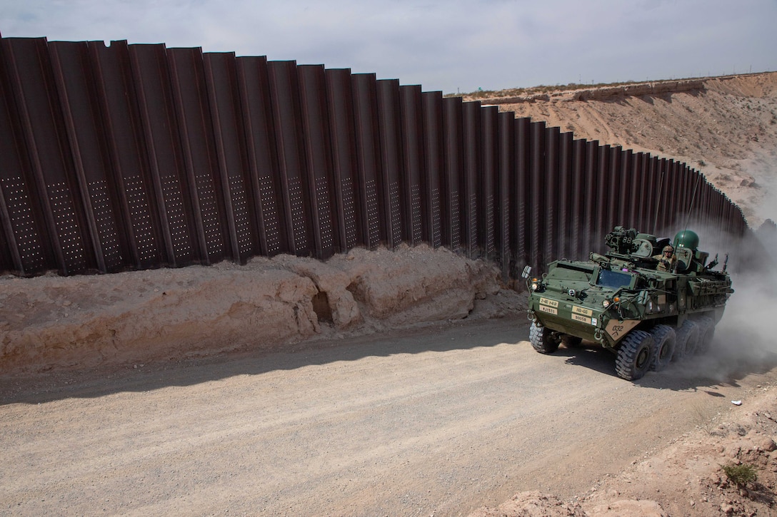 Soldiers drive armored military vehicle on a dusty road next to a tall fence.