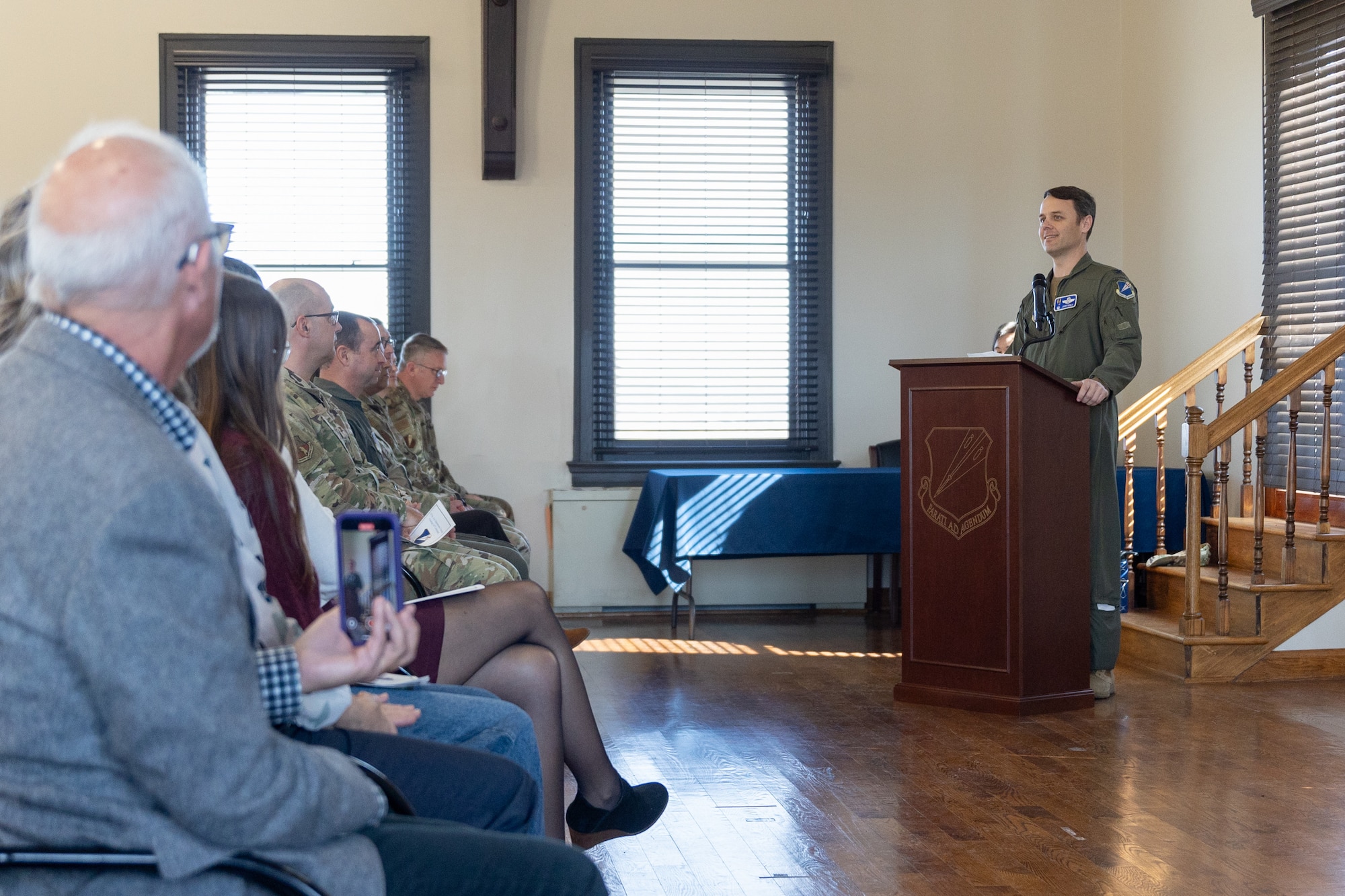 An Air National Guard Airman speaks at a podium to a crowd of military members and civilians. The Airman is wearing a military flight suit.