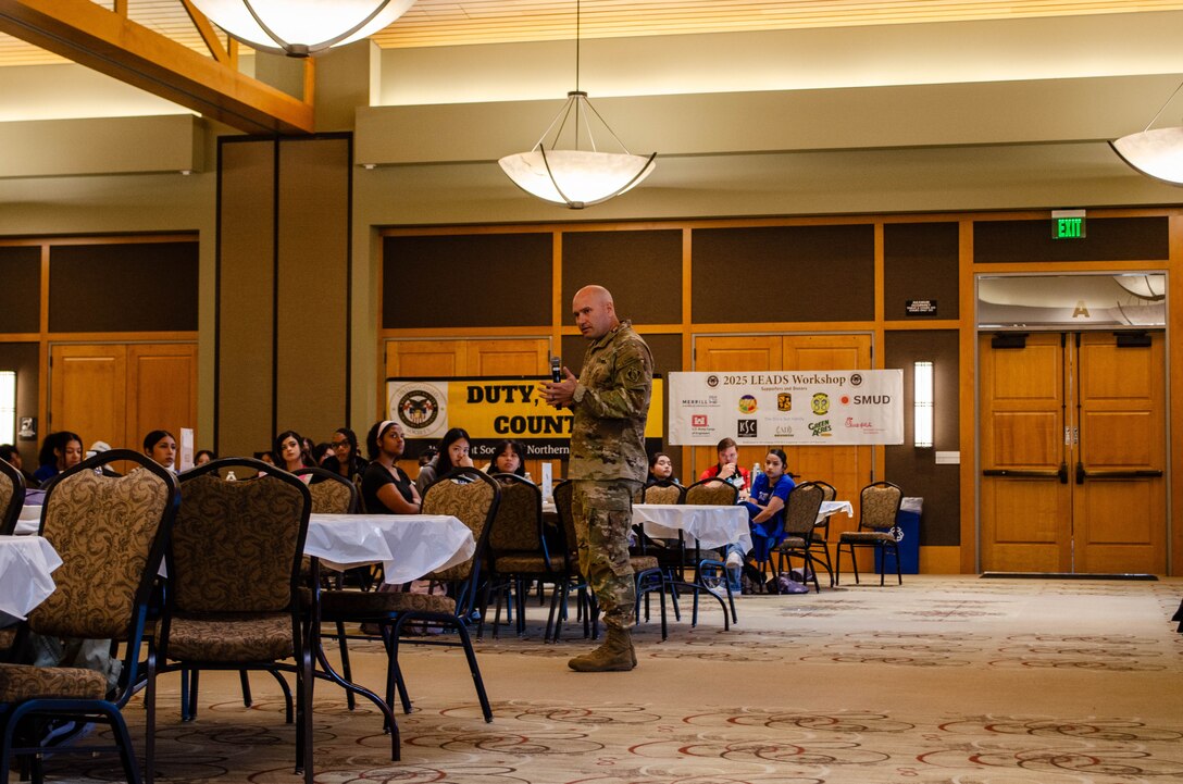 USACE Sacramento District Commander Col. Chad Caldwell stands in ballroom with round tables and chairs to give a presentation to students about STEM careers.