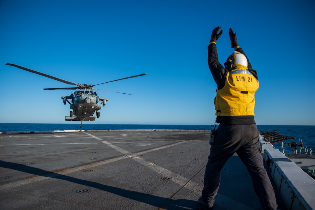 A sailor wearing a yellow vest signals to a helicopter landing on the flight deck of a military ship.