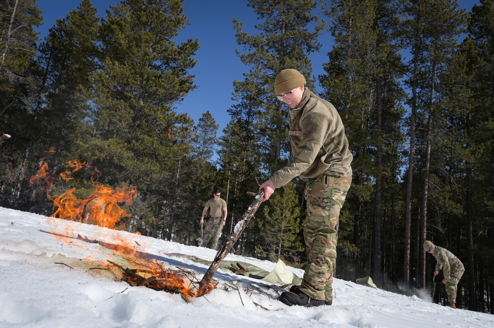 A person in military uniform uses a stick to cover a small campfire in snow.