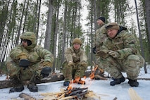 Three people in military uniform crouch around a small campfire in the snow.