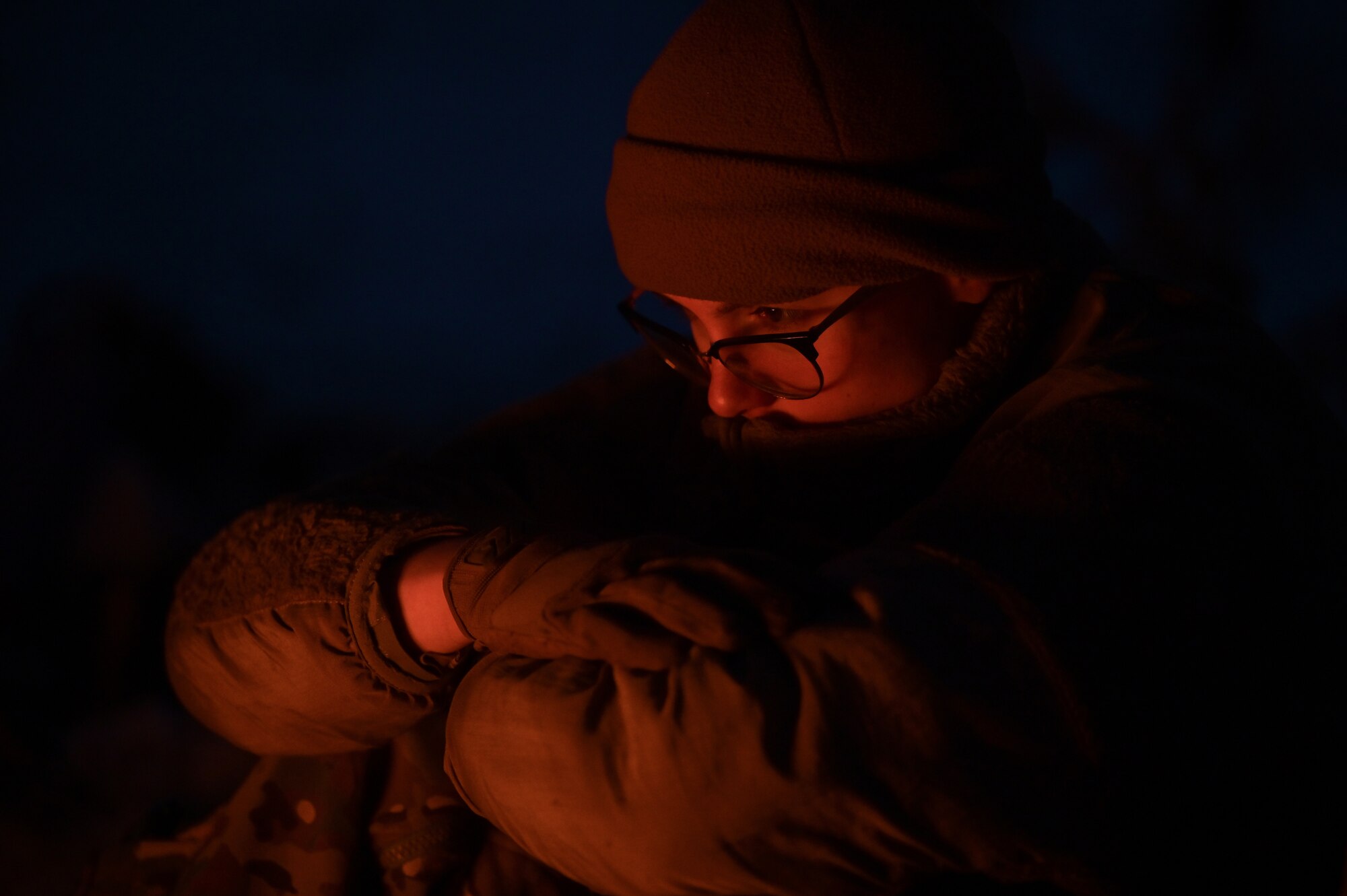 A person's face is slightly illuminated in an orange flow from a campfire in front of them.