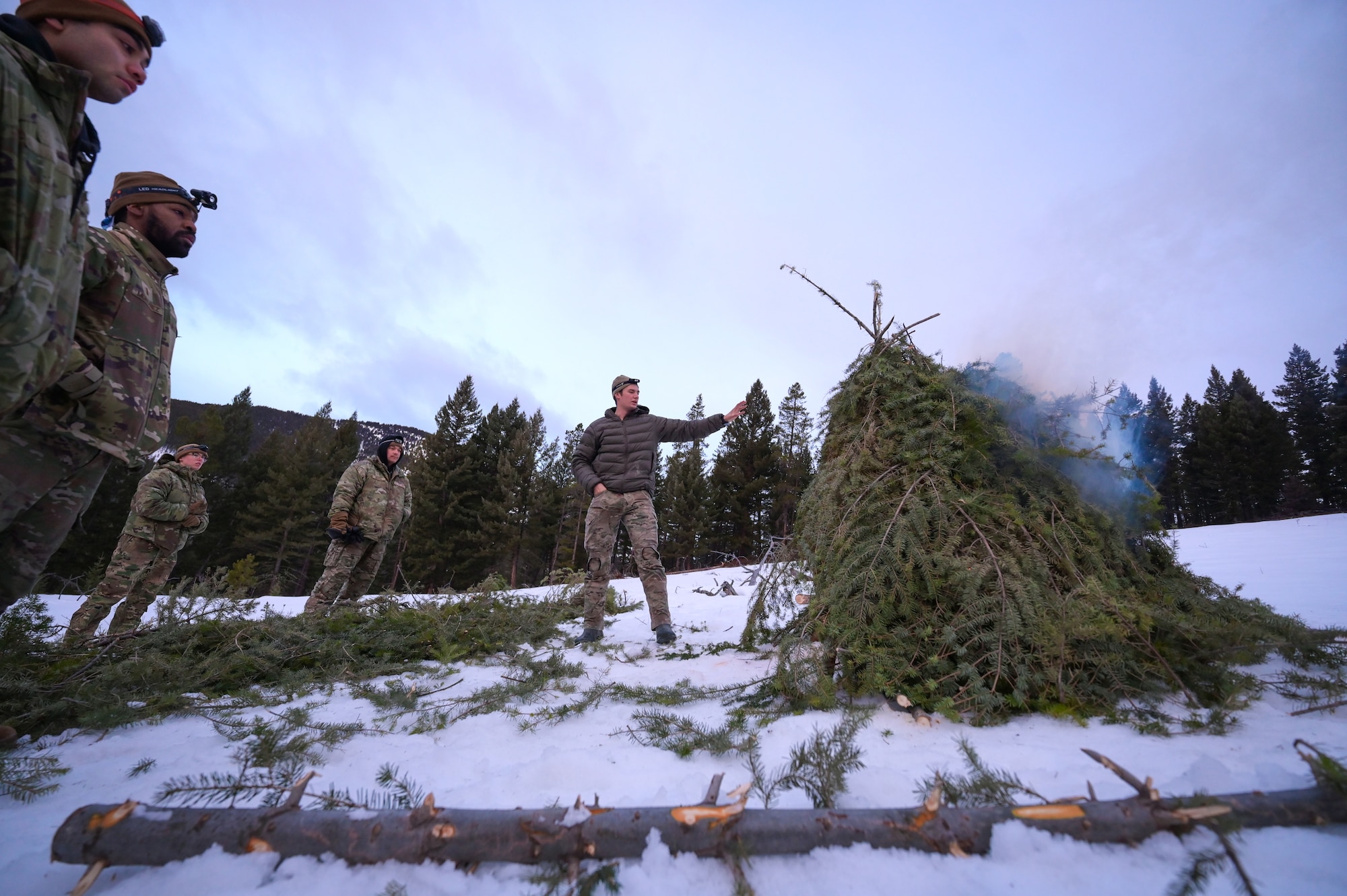 A group of people observe a pyre of tree boughs as it begins to smoke.