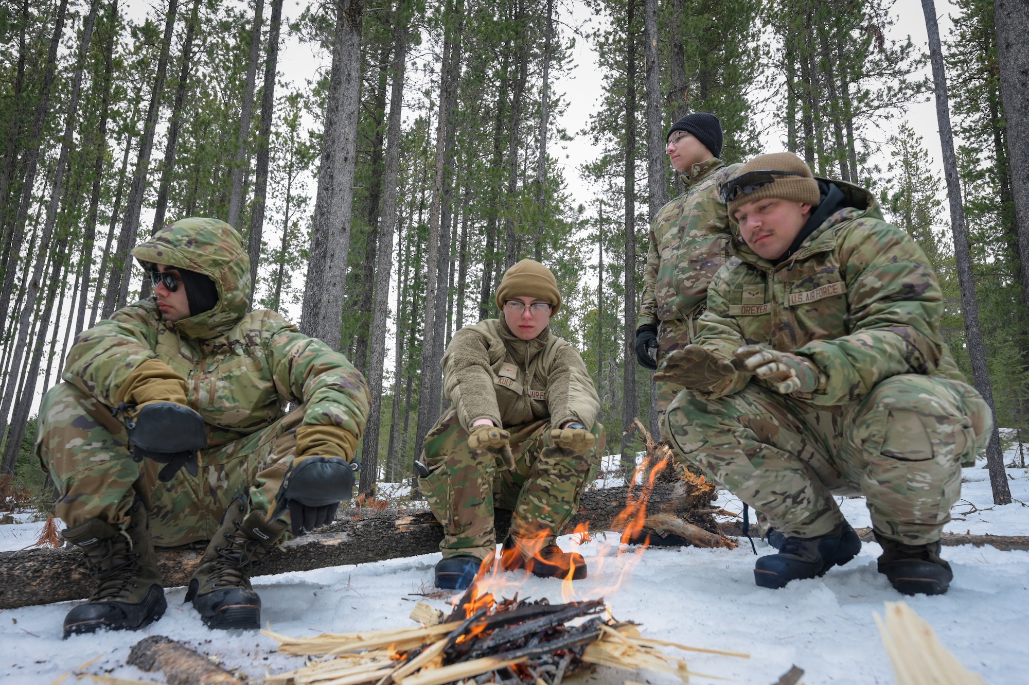 Three people in military uniform crouch around a small campfire in the snow.