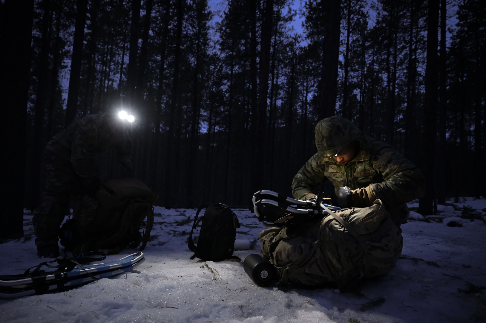 A person in military uniform crouches in the dark to pack their rucksack under the glow of a headlamp.