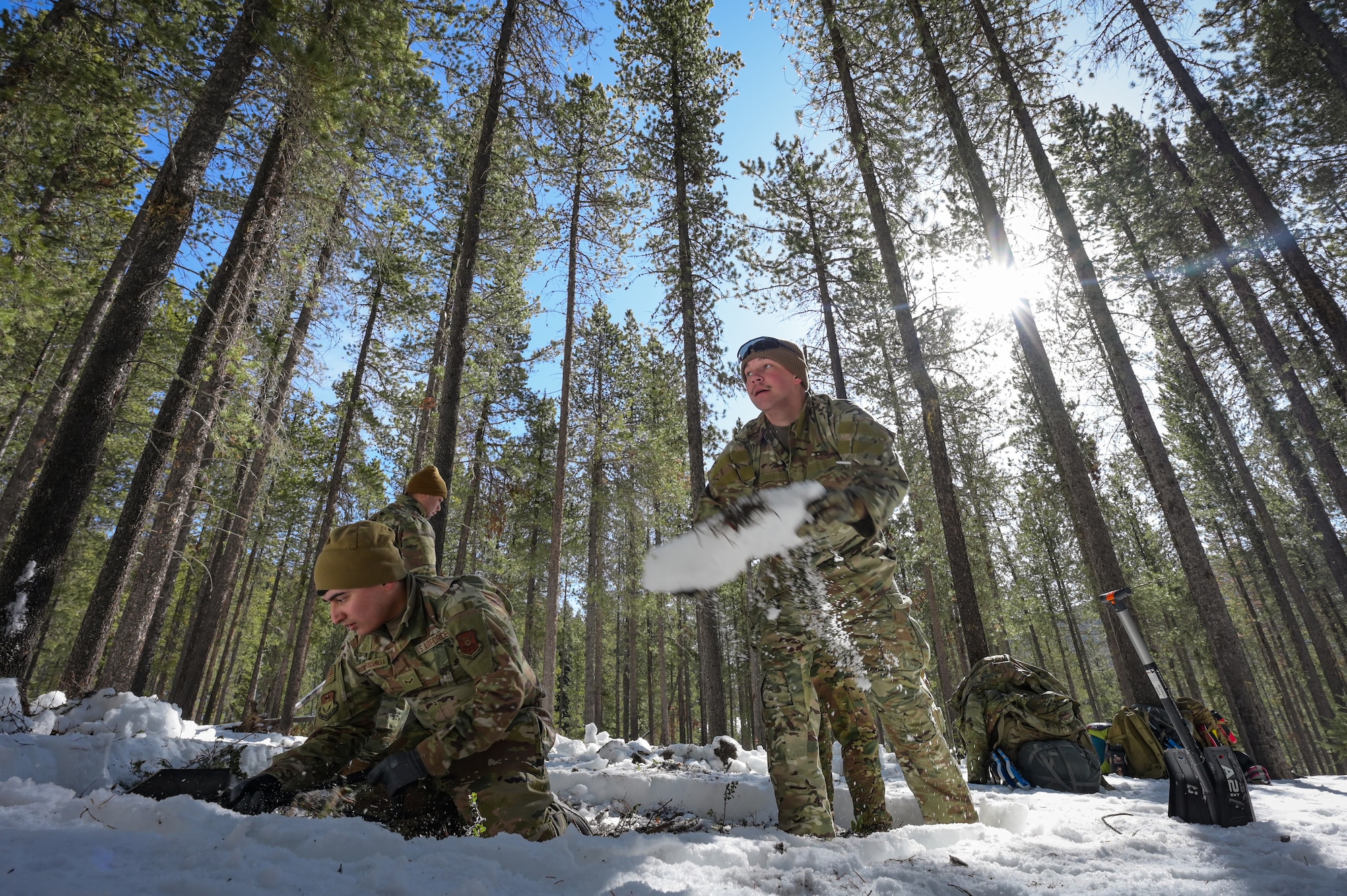 Two men in military uniform work together to dig in the snow to create a fire circle.