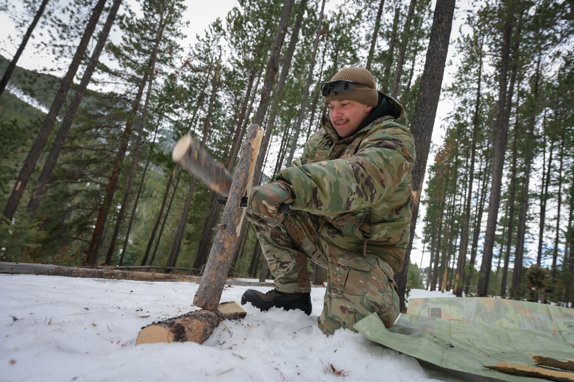 A man in military uniform splits firewood using a fixed blade knife and batoning technique.