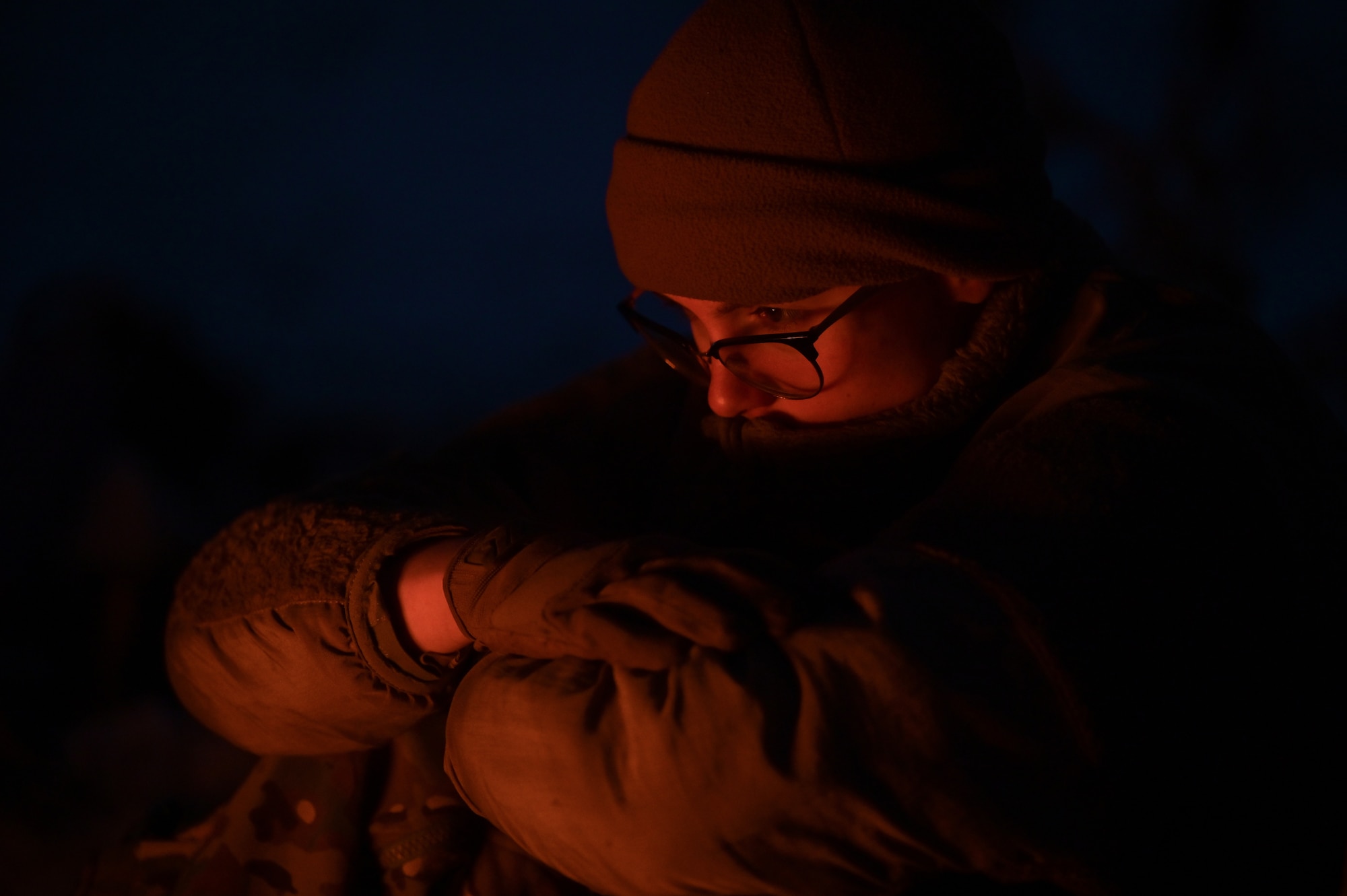 A person's face is slightly illuminated in an orange flow from a campfire in front of them.