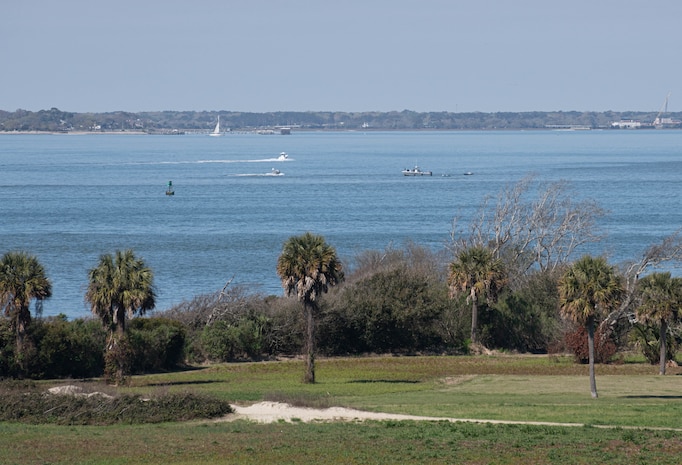Photo overview of Sullivan's Island with palmetto trees and Charleston bay in the background with naval assets.