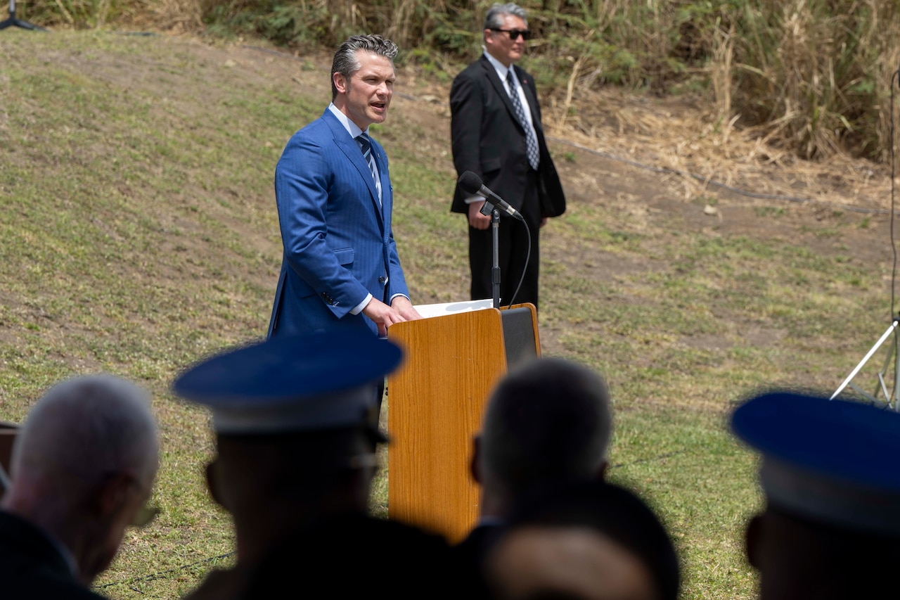 Defense Secretary Pete Hegseth stands and speaks into a mic at a lectern with another person standing to the left and others seated in the foreground.